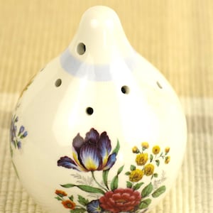 Puede incluir: Salero de cerámica blanca con forma bulbosa y varios agujeros en la parte superior. Está decorado con un diseño floral con flores moradas, rojas, amarillas y azules. Una franja azul claro rodea la parte superior.