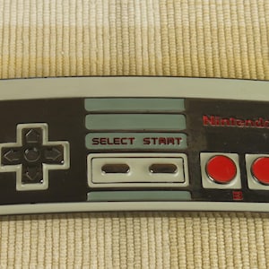May include: A rectangular, retro-style Nintendo controller with a grey border. The controller features a black directional pad, grey and teal accents, red buttons, and the text "SELECT START" and "Nintendo".