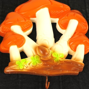 May include: A whimsical wall hook featuring a cluster of orange and white mushrooms with a brown base and a single hook. The design includes green shamrocks and a small acorn, creating a nature-inspired decorative piece.