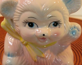 May include: A pink ceramic piggy bank shaped like a bear with blue eyes, a yellow bow, and blue accents on the ears and feet.