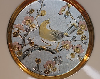 Könnte beinhalten: Ein dekorativer Teller mit goldfarbenem Rand und silberner Mitte, der ein zartes Blumenmuster mit einem kleinen gelben Vogel auf einem Zweig zeigt. Die Blumen sind rosa, weiß und goldfarben.