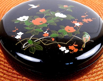May include: A black round box with a floral design. The box is decorated with white, red, and green flowers and a small bird. The box is sitting on an orange surface.