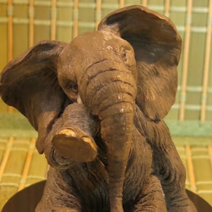 May include: A small, brown, ceramic elephant figurine with a textured surface. The elephant is sitting with its trunk raised and its ears folded back.