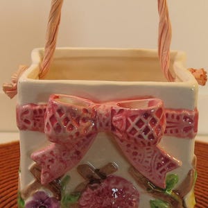 May include: A white ceramic planter shaped like a basket with a pink ribbon and floral details. The planter has a twisted rope handle and features pink, yellow, and purple flowers.