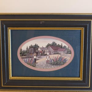 以下が含まれることがあります： 馬車、家、人物が描かれた田園風景を描いた額装されたアートワーク。 楕円形の画像は、濃いティール色の背景に配置され、金と黒で縁取られています。 アートワークはピンク色のボーダーと穏やかな風景が特徴です。