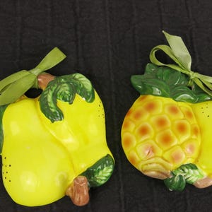 May include: Two ceramic fruit ornaments: a yellow pear and a pineapple. The pear has green leaves and a green ribbon. The pineapple is yellow with brown accents and green leaves. Both have a glossy finish.