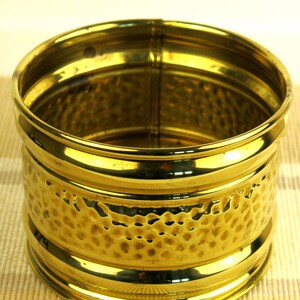 May include: A cylindrical brass container with a hammered texture on the middle section. The container has a shiny, reflective surface and a simple, elegant design. The container is suitable for holding small items.
