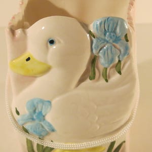 May include: A white ceramic planter shaped like a duck with blue and yellow flowers. The planter has a white rope handle and a pink and white checked pattern on the inside.