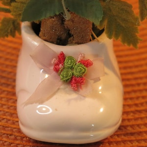 Peut inclure: Un pot de fleurs en forme de chaussure blanche en céramique avec un ruban rose et un motif floral. Le pot est rempli d'une petite plante verte.