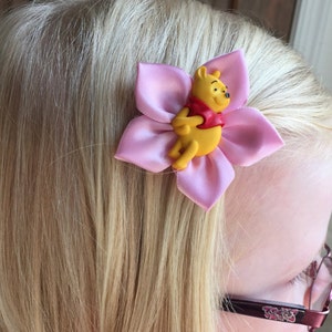May include: A pink flower hair clip with a yellow Winnie the Pooh character in the center.