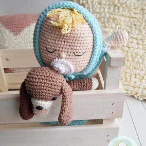 Puede incluir: Una mu&ntilde;eca de crochet con un chupete, que lleva un atuendo azul con capucha, est&aacute; sosteniendo un cachorro de crochet marr&oacute;n. La mu&ntilde;eca y el cachorro est&aacute;n en una caja de madera blanca.