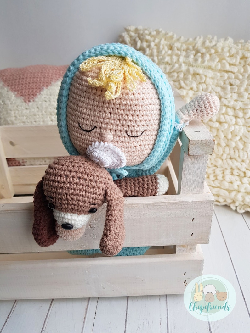 Puede incluir: Una mu&ntilde;eca de crochet con un chupete, que lleva un atuendo azul con capucha, est&aacute; sosteniendo un cachorro de crochet marr&oacute;n. La mu&ntilde;eca y el cachorro est&aacute;n en una caja de madera blanca.