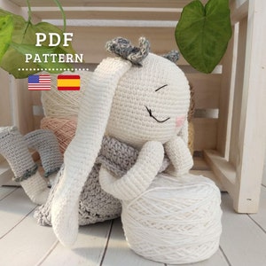 Op de afbeelding: Een gehaakt amigurumi-speelgoed in de vorm van een konijn met een wit lichaam, een grijze jurk en een witte bol wol. Het konijn heeft een expressie met gesloten ogen en een kleine roze neus. Het speelgoed zit op een houten oppervlak. De tekst "PDF PATTERN" is zichtbaar in de afbeelding.