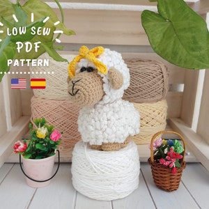 Könnte beinhalten: Ein gehäkeltes weißes Schaf-Amigurumi-Spielzeug mit einer gelben Schleife, das auf einer weißen Wollkugel sitzt. Das Schaf steht vor einem Holzregal mit Wollknäueln in beige und cremefarben. Das Bild zeigt auch zwei kleine Blumenarrangements in Töpfen.