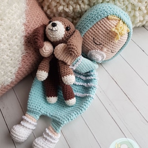 Puede incluir: Una mu&ntilde;eca de beb&eacute; de crochet con los ojos cerrados, vestida con un atuendo azul claro y sosteniendo un cachorro de crochet marr&oacute;n y blanco. La mu&ntilde;eca est&aacute; tumbada sobre una superficie de madera blanca.