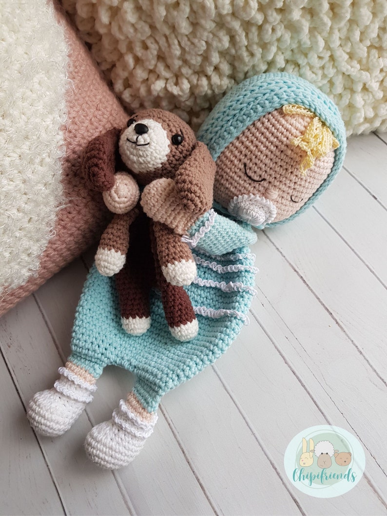 Puede incluir: Una mu&ntilde;eca de beb&eacute; de crochet con los ojos cerrados, vestida con un atuendo azul claro y sosteniendo un cachorro de crochet marr&oacute;n y blanco. La mu&ntilde;eca est&aacute; tumbada sobre una superficie de madera blanca.