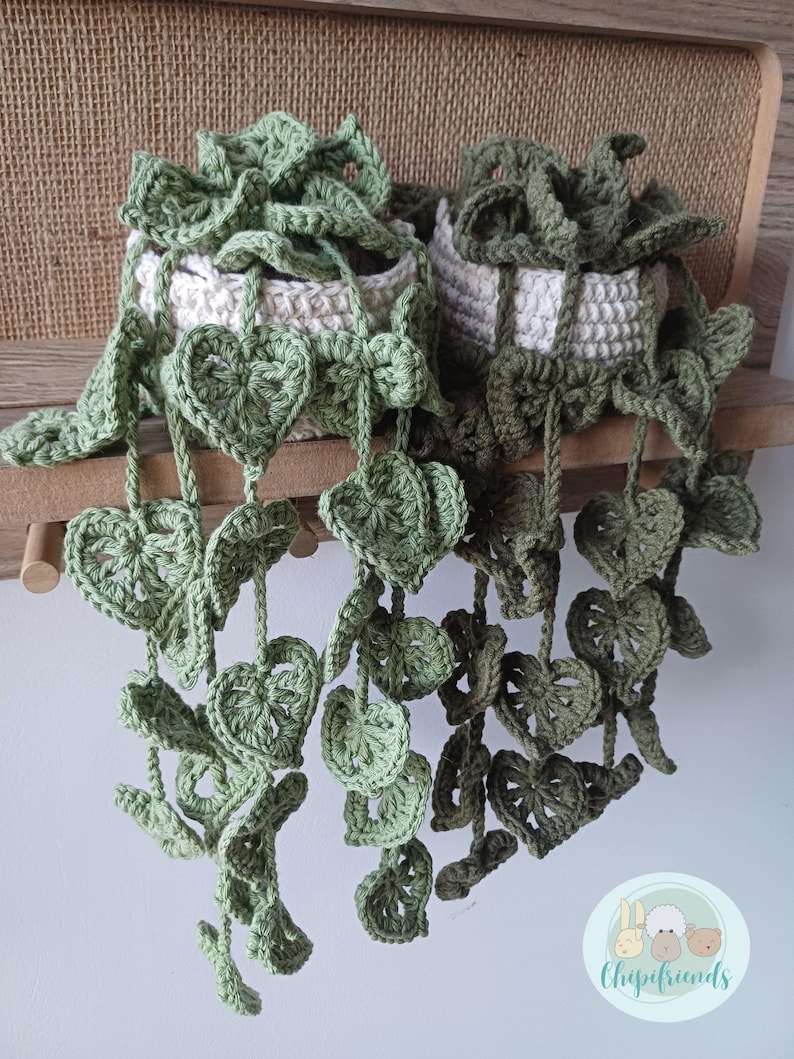 May include: Two crocheted hanging plants in white pots with green leaves. The plants are hanging from a wooden shelf. The leaves are in the shape of hearts. The plants are made by Chipifriends.
