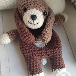 Puede incluir: Un juguete de perro de crochet marr&oacute;n con detalles blancos. El perro tiene una cara blanca con ojos negros y una nariz negra. El perro est&aacute; sentado con las patas metidas.