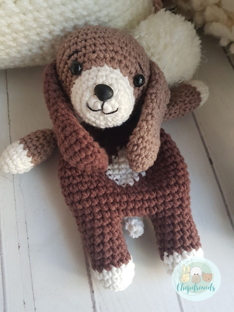 Puede incluir: Un juguete de perro de crochet marr&oacute;n con detalles blancos. El perro tiene una cara blanca con ojos negros y una nariz negra. El perro est&aacute; sentado con las patas metidas.
