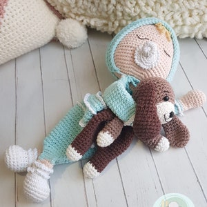 Puede incluir: Una mu&ntilde;eca de crochet con un chupete y un cachorro de crochet marr&oacute;n. La mu&ntilde;eca lleva un atuendo azul claro y blanco y un lazo amarillo en el pelo. La mu&ntilde;eca y el cachorro est&aacute;n tumbados sobre una superficie de madera blanca.