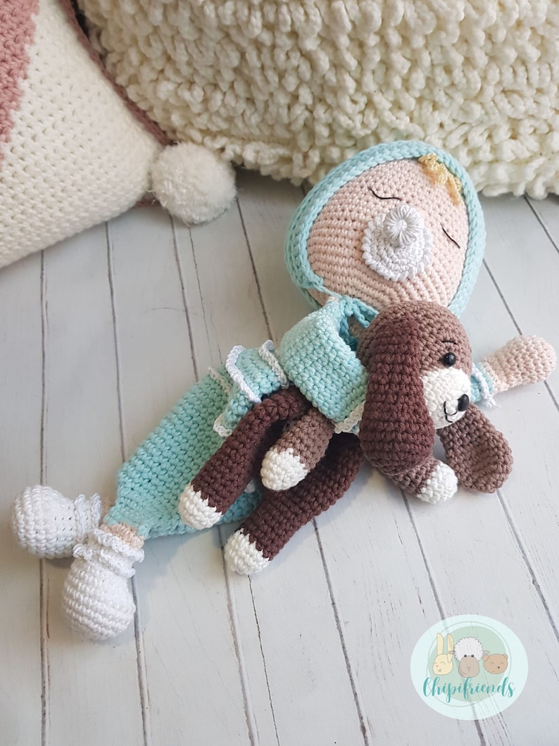 Puede incluir: Una mu&ntilde;eca de crochet con un chupete y un cachorro de crochet marr&oacute;n. La mu&ntilde;eca lleva un atuendo azul claro y blanco y un lazo amarillo en el pelo. La mu&ntilde;eca y el cachorro est&aacute;n tumbados sobre una superficie de madera blanca.