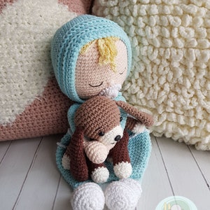 Puede incluir: Una mu&ntilde;eca de crochet con cabello rubio, que lleva un gorro y un atuendo azul y blanco, sostiene un cachorro de crochet marr&oacute;n. La mu&ntilde;eca est&aacute; acostada sobre una almohada de crochet blanca y marr&oacute;n.
