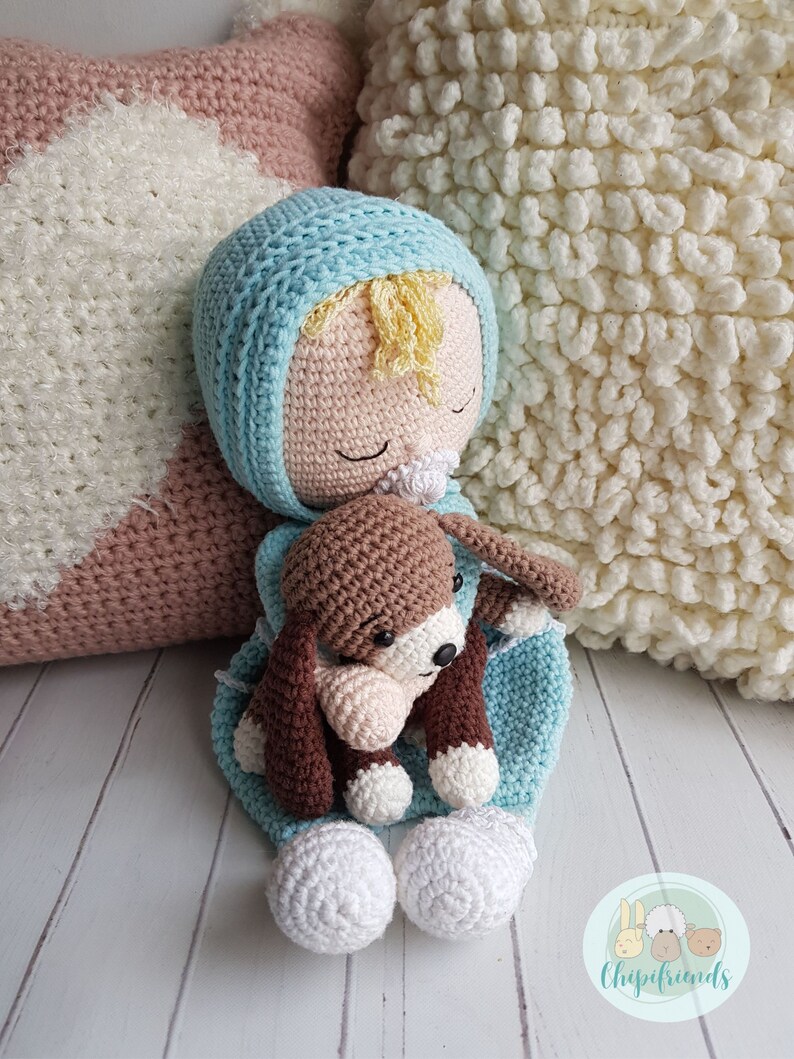 Puede incluir: Una mu&ntilde;eca de crochet con cabello rubio, que lleva un gorro y un atuendo azul y blanco, sostiene un cachorro de crochet marr&oacute;n. La mu&ntilde;eca est&aacute; acostada sobre una almohada de crochet blanca y marr&oacute;n.