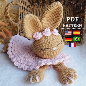 Pode incluir: Um coelho de crochê vestindo um vestido rosa com uma coroa de flores. O coelho está deitado em uma superfície branca. A imagem inclui o texto "PDF PATTERN" e bandeiras dos Estados Unidos, Espanha, Alemanha, Brasil e França.