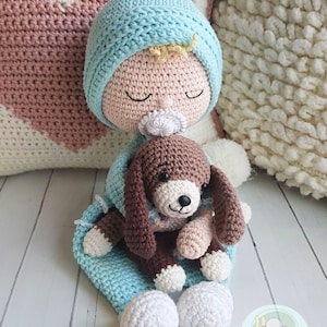 Puede incluir: Una mu&ntilde;eca de crochet beb&eacute; con un gorro azul y un cachorro de crochet marr&oacute;n en sus brazos. La mu&ntilde;eca lleva un chupete blanco y tiene los ojos cerrados. La mu&ntilde;eca est&aacute; sentada sobre una superficie blanca con una manta rosa y blanca detr&aacute;s.