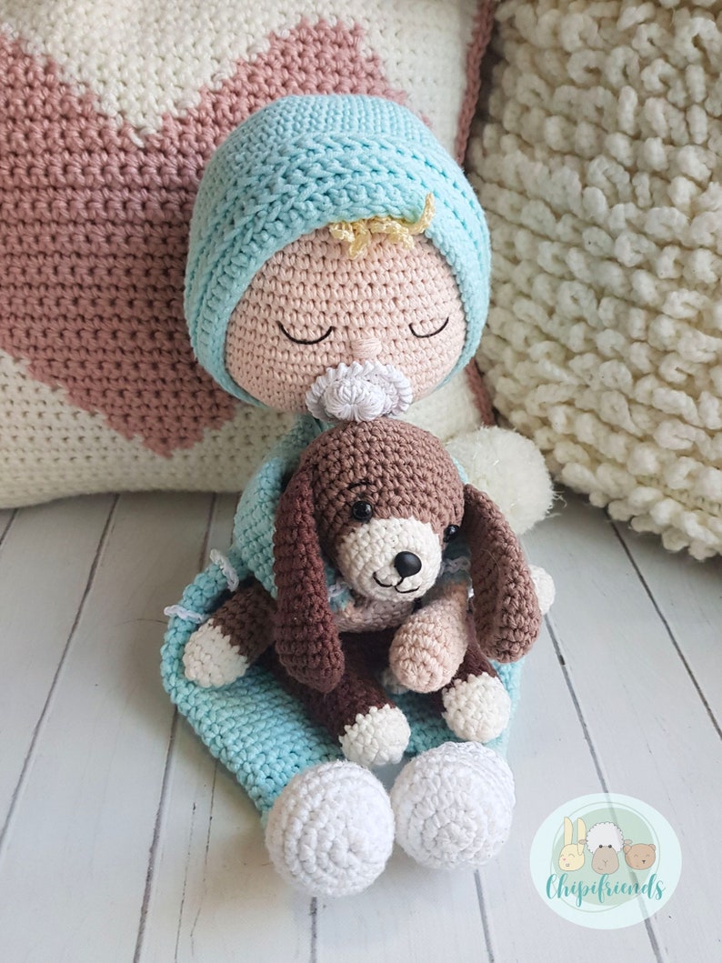 Puede incluir: Una mu&ntilde;eca de crochet beb&eacute; con un gorro azul y un cachorro de crochet marr&oacute;n en sus brazos. La mu&ntilde;eca lleva un chupete blanco y tiene los ojos cerrados. La mu&ntilde;eca est&aacute; sentada sobre una superficie blanca con una manta rosa y blanca detr&aacute;s.