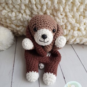 Puede incluir: Un juguete de perro de crochet marr&oacute;n y blanco con una bufanda marr&oacute;n. El juguete est&aacute; sentado sobre una superficie de madera blanca.