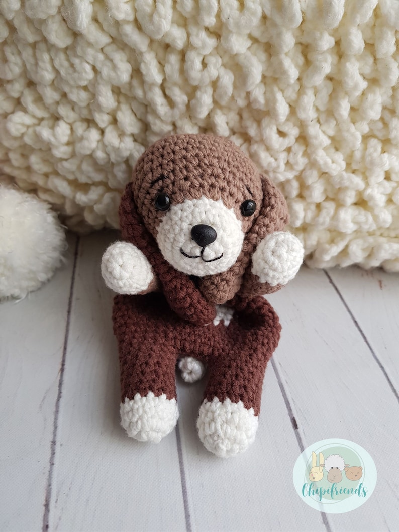 Puede incluir: Un juguete de perro de crochet marr&oacute;n y blanco con una bufanda marr&oacute;n. El juguete est&aacute; sentado sobre una superficie de madera blanca.