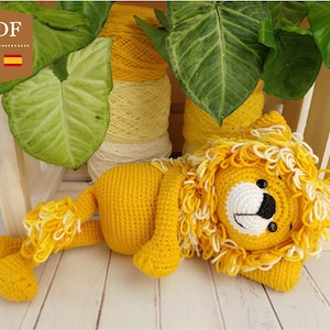Puede incluir: Un león de crochet amarillo con una melena blanca, acostado sobre una superficie de madera. El león tiene ojos negros y una nariz negra.
