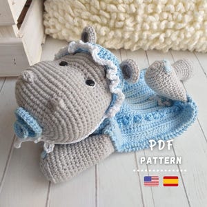 Könnte beinhalten: Ein gehäkeltes graues Flusspferd, das ein blaues und weißes gehäkeltes Kleid trägt. Das Flusspferd liegt auf einer weißen Holzoberfläche. Der Text "PDF PATTERN" ist auf dem Bild zu sehen. Die amerikanische und spanische Flagge sind ebenfalls auf dem Bild zu sehen.