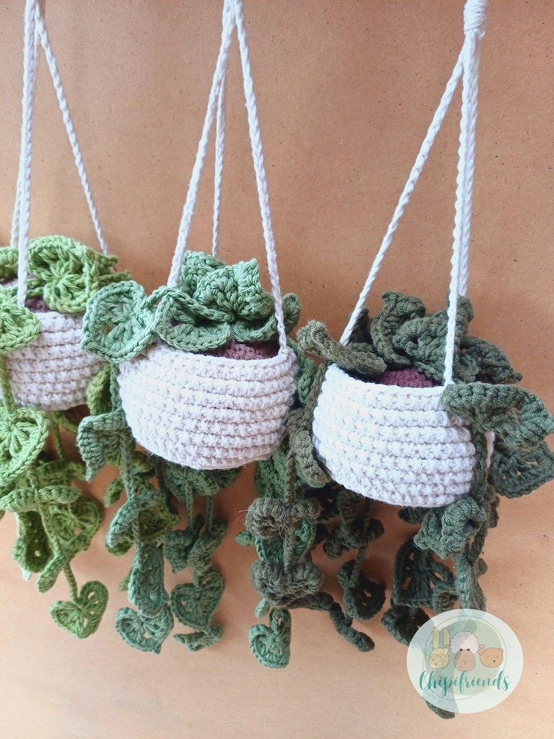May include: Three crocheted hanging planters with green leafy plants. The planters are made of a beige yarn and have a white cord for hanging.