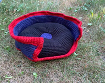 Puede incluir: Una cama para mascotas hecha a mano con ganchillo en una combinación de colores vibrantes. La cama presenta un interior negro, una sección media azul y un borde exterior rojo. La cama es redonda y está sobre hierba verde.
