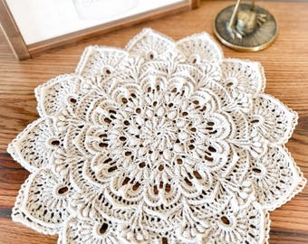 Puede incluir: Tapete de ganchillo hecho a mano de color crema con un diseño floral. El tapete presenta un patrón calado detallado con bordes festoneados. El intrincado diseño está centrado en una superficie de madera, mostrando la artesanía de la pieza.