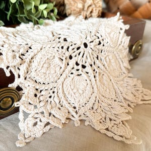 May include: A cream-coloured, handmade crochet doily with a floral design. The doily features a central flower motif surrounded by repeating leaf-like patterns. The edges are scalloped, adding a delicate touch.
