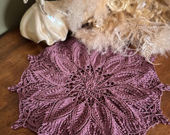 Puede incluir: Un tapete de ganchillo circular de color malva con un intrincado patrón radiante en forma de hoja con puntadas texturizadas. El diseño incluye un punto central que se expande hacia afuera en bordes puntiagudos y detallados de tejido artesanal.