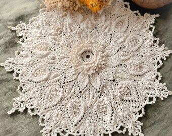 Puede incluir: Tapete de ganchillo hecho a mano de color crema con un diseño floral. El tapete presenta una flor central y patrones de hojas intrincados, con un borde festoneado. El tapete se muestra sobre una superficie verde, con flores secas y un objeto decorativo en el fondo.