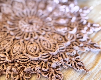 Puede incluir: Un delicado tapete de ganchillo hecho a mano en un suave color malva. El intrincado diseño presenta un patrón floral central con múltiples capas de bordes texturizados. El tapete se muestra sobre una superficie clara y texturizada.