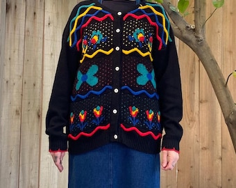 May include: A black cardigan sweater with colorful floral and abstract patterns. The sweater has a button-up front and is paired with a long denim skirt. The sweater's design includes red, yellow, blue, and green accents.