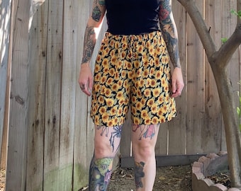 Puede incluir: Pantalones cortos a medio muslo con un estampado de girasoles de pétalos amarillos y centros marrones sobre un fondo negro. Los pantalones cortos tienen una cintura elástica con un cordón ajustable a juego en el centro delantero.