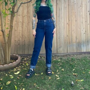 Op de afbeelding: Een persoon draagt een zwarte top en een donkerblauwe jeans. De jeans heeft een rechte pijp en is omgeslagen bij de enkels. De persoon draagt ook zwarte sandalen. De achtergrond is een houten hek en groen gras.