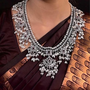 May include: A silver-toned necklace and matching earrings set. The necklace features multiple layers with intricate floral and geometric designs, accented with small white pearls and red gemstones. The earrings have a similar design, with dangling pearl accents.