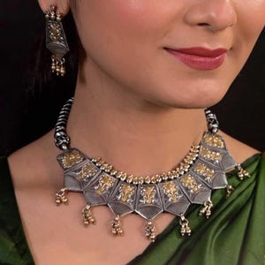 May include: A silver and gold necklace with a geometric design and dangling charms. The necklace is worn by a person with dark hair and a green shirt.