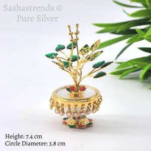 May include: A small, silver Tulsi plant figurine with green leaves and red flowers. The plant is in a gold pot with a white base and pink and white embellishments. The figurine is 7.4 cm tall and the base is 3.8 cm in diameter.