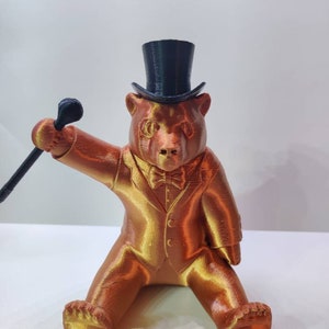 May include: A 3D printed figurine of a bear wearing a black top hat, a bow tie, and a suit. The bear is holding a cane in its right paw and is sitting with its legs crossed.