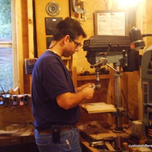 Pu&ograve; includere: Una persona che lavora in un laboratorio di falegnameria, usando una trapano a colonna. Il laboratorio ha pareti in legno e una finestra. &Egrave; visibile una sega a nastro Delta 9". L'indirizzo email customshifters@gmail.com &egrave; visibile.