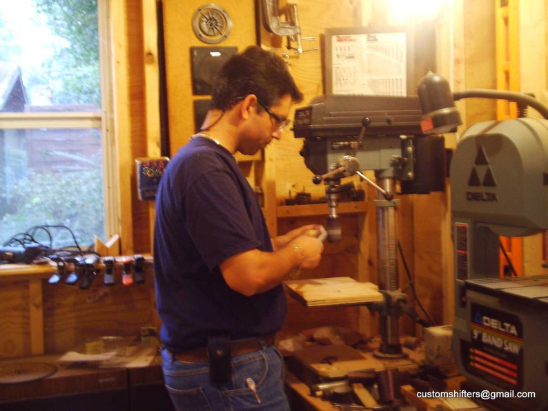 Pu&ograve; includere: Una persona che lavora in un laboratorio di falegnameria, usando una trapano a colonna. Il laboratorio ha pareti in legno e una finestra. &Egrave; visibile una sega a nastro Delta 9". L'indirizzo email customshifters@gmail.com &egrave; visibile.